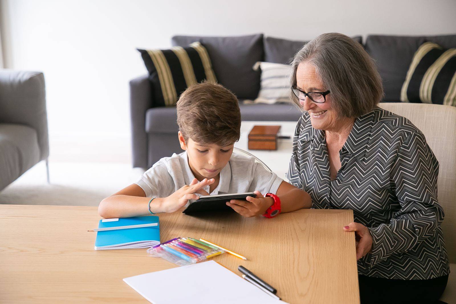 Une grand-mère penchée vers un enfant qui utilise une tablette sur une table basse.